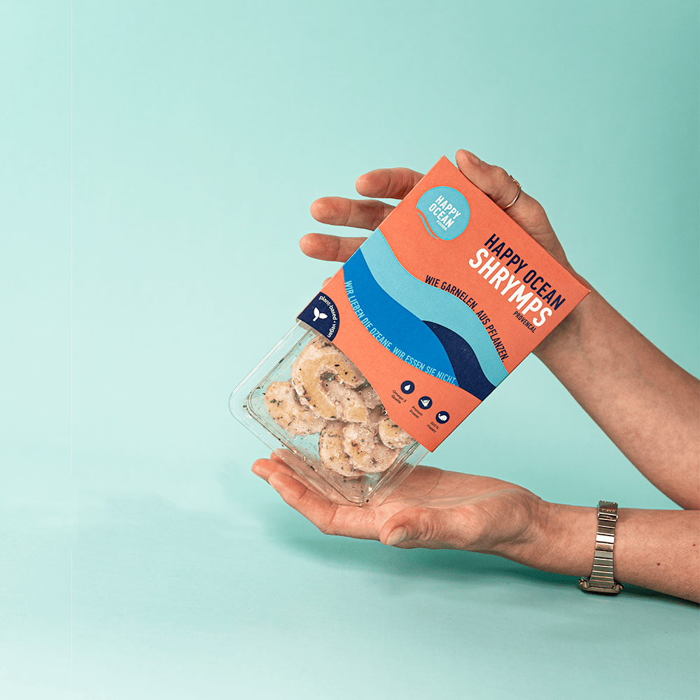 two hands reaching into the image from the right holding a package of Happy Ocean Shrymps in a vibrant orange/pink packaging'