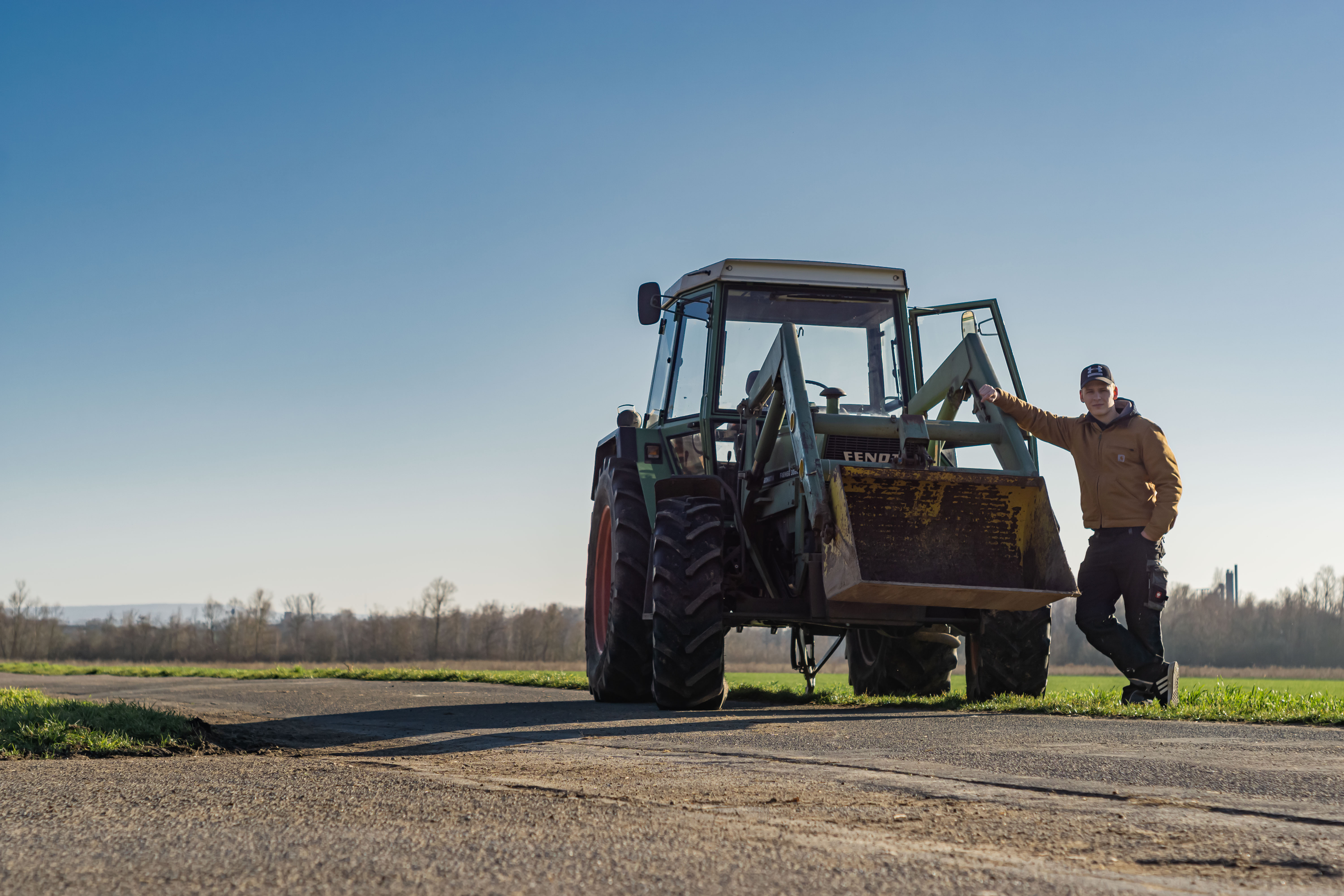 young man in engelbert strauss working clothes standing infront of a tractor, which is parked infront of a field