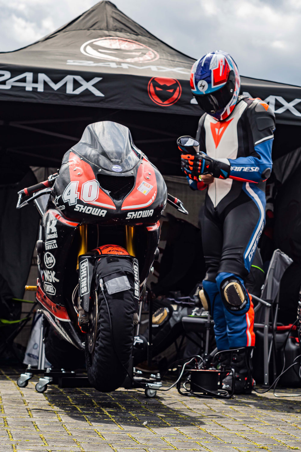 man in a colorful red, white, black and blue leathersuite standing next to his racing motorbike. Tent in the background