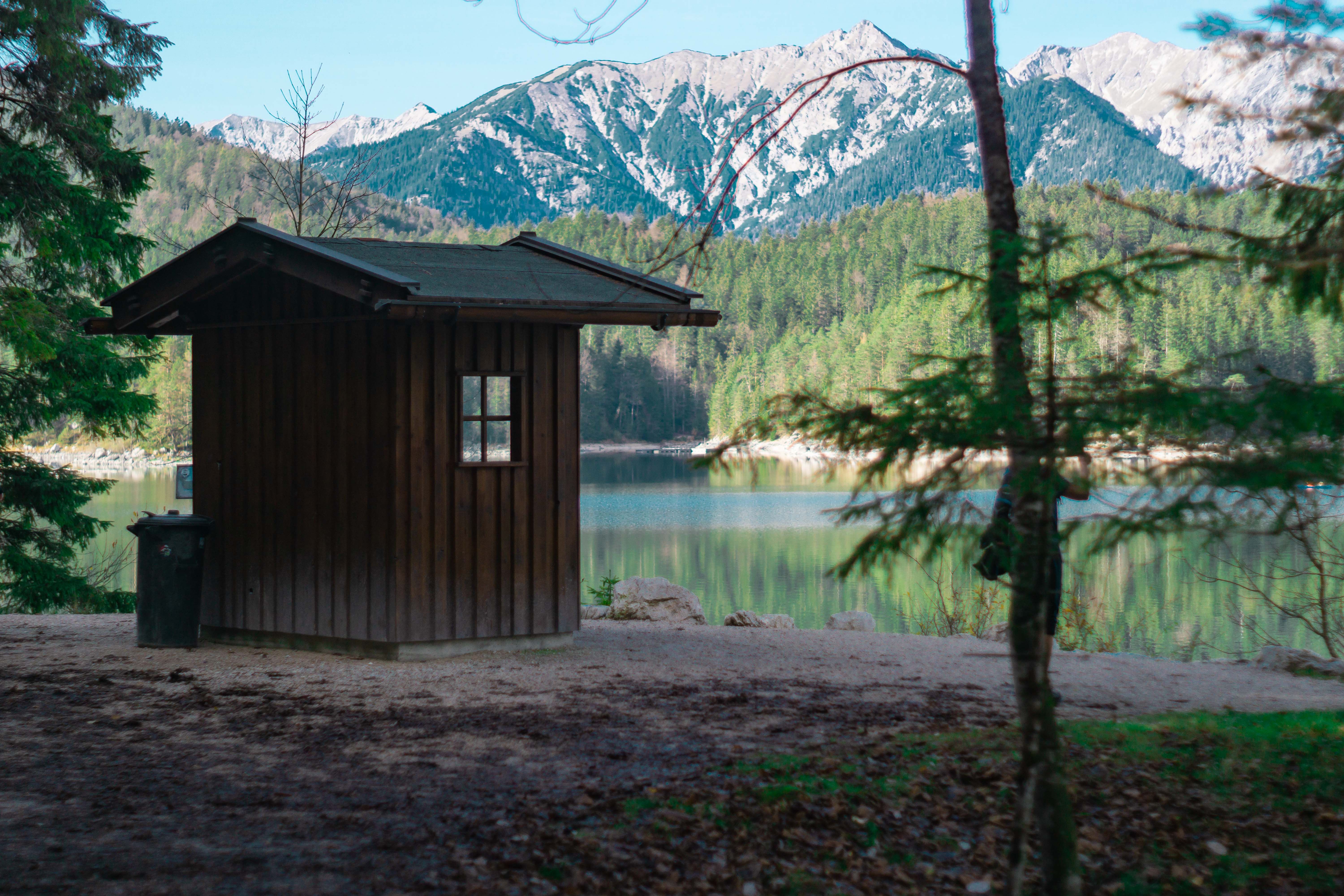 Photograph of a small forest hut. Photographed from behind. The front of the hut is facing the lake, behind which a green coniferous forest and high mountains can be seen against a blue sky.