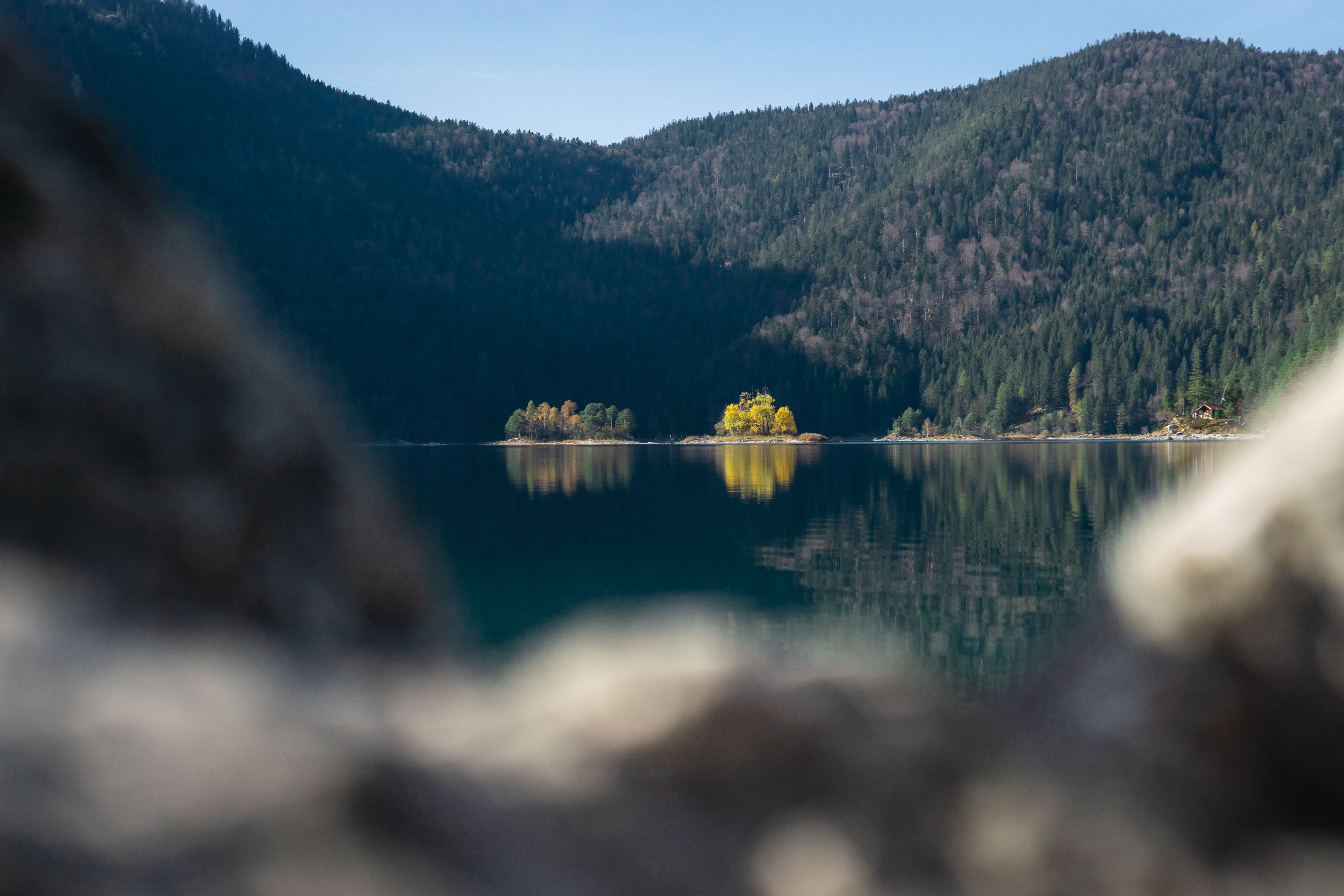 Photograph of a lake: in the foreground are blurred rough stones, in the focus iare two small round islands with autumnal yellow trees on a lake in the far distance. Mountains can be seen in the background.