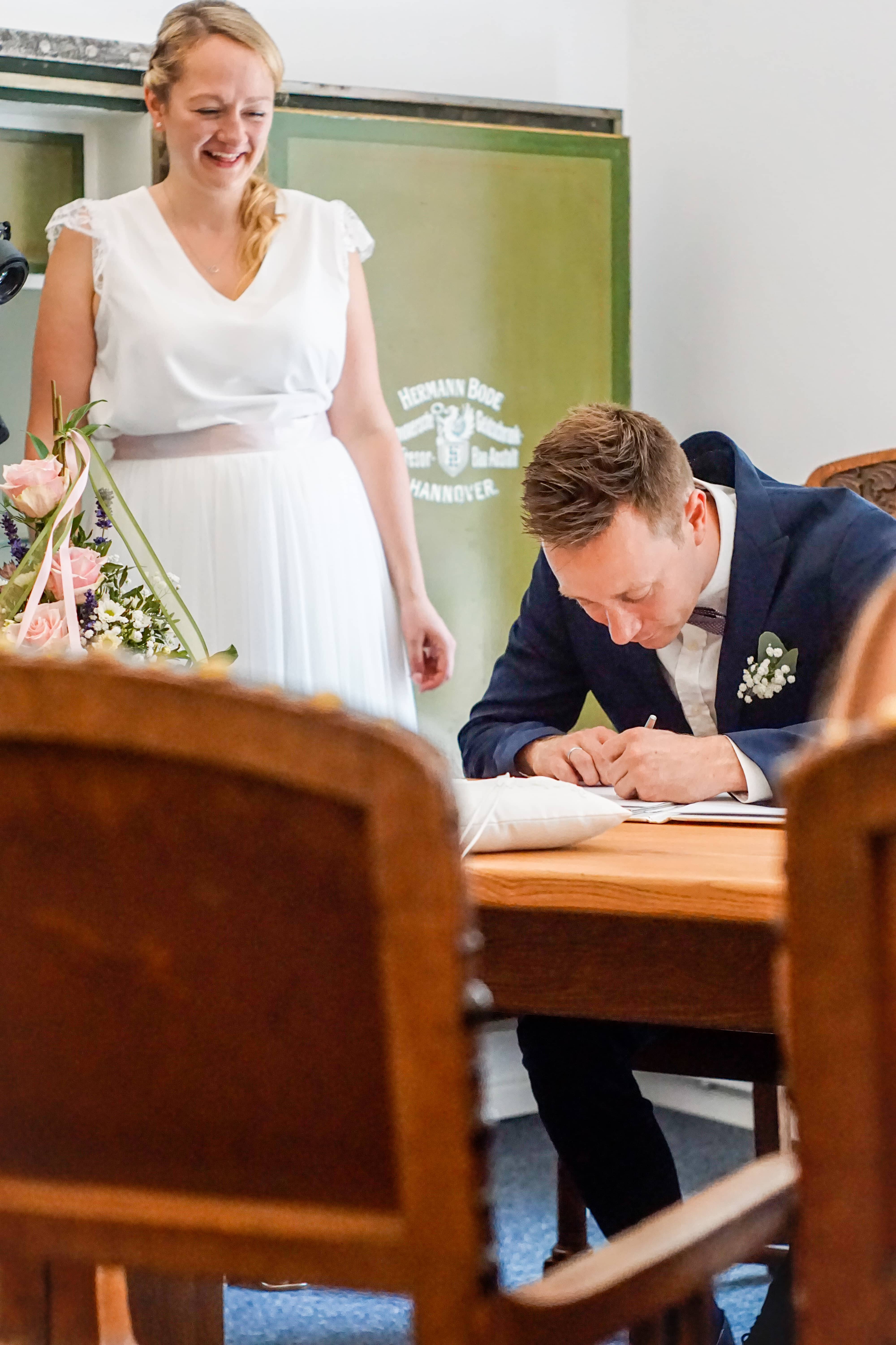 groom signing the marriage document while the bride grins jokily behind him